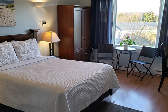 Slaapkamer met een wit bed, lamp, kledingkast en een klein tafeltje bij een zonnig raam.