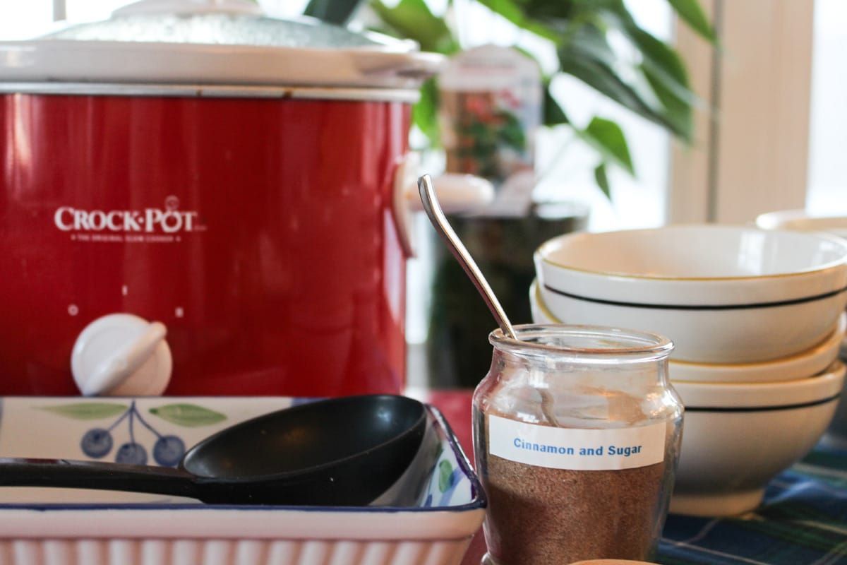 Een rode slowcooker staat op een tafel naast kommen, een lepel en een potje saus.