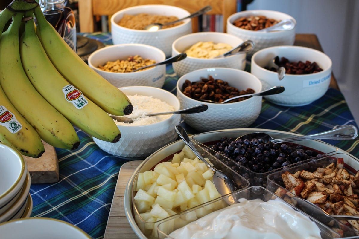 Bananen en schalen met fruit en diverse toppings staan uitgestald op een tafelkleed voor een ontbijtbuffet.