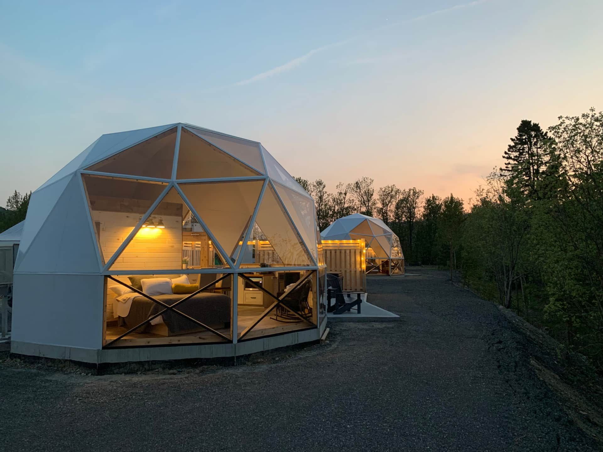 Geodetische koepelhutten van Appalaches Domes die in de schemering oplichten langs een grindpad naast bomen.