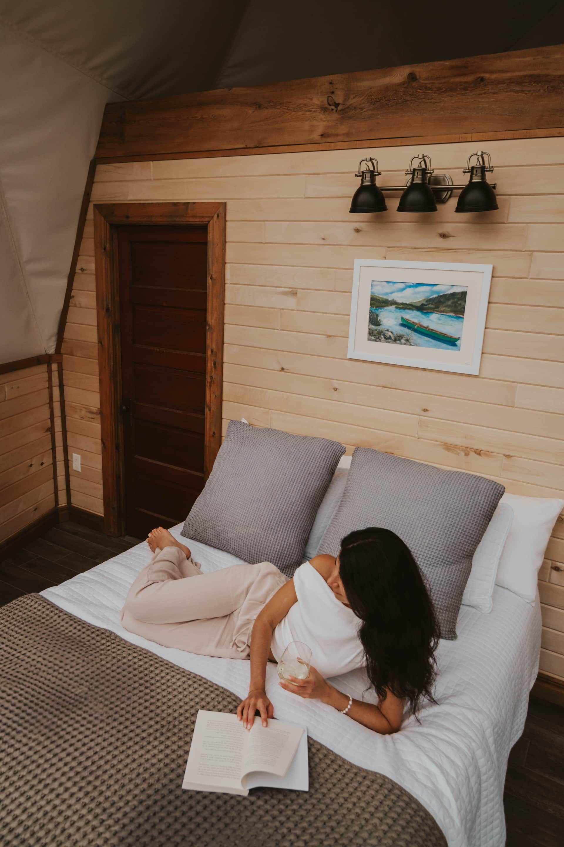 Een persoon leest op een bed in een gezellige houten slaapkamer met grijze kussens en wanddecoratie.
