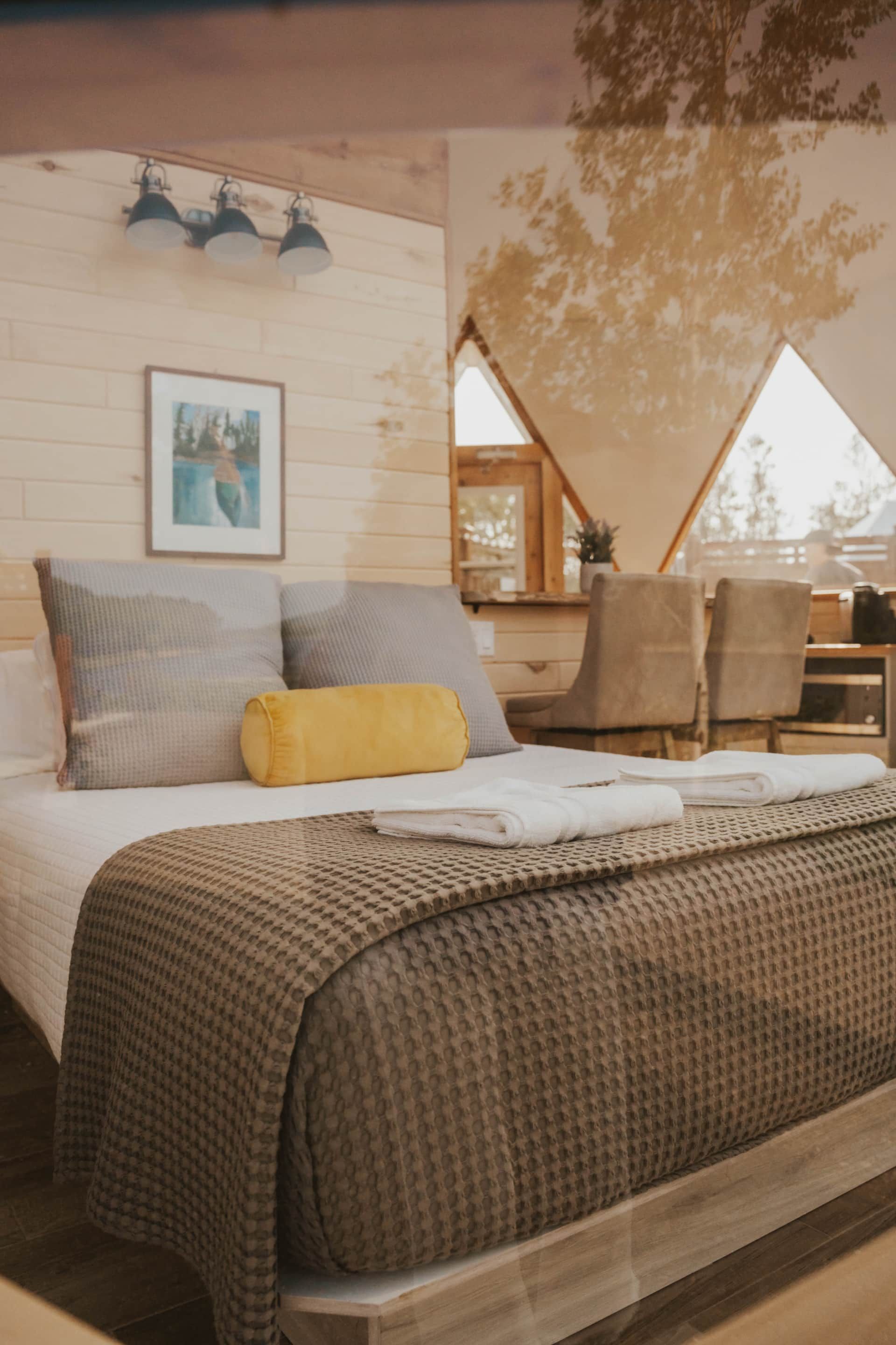 Gezellige slaapkamer met een bed, gestreepte kussens, een geel sierkussen, ingelijste kunst en een houten accentmuur met driehoekige ramen.