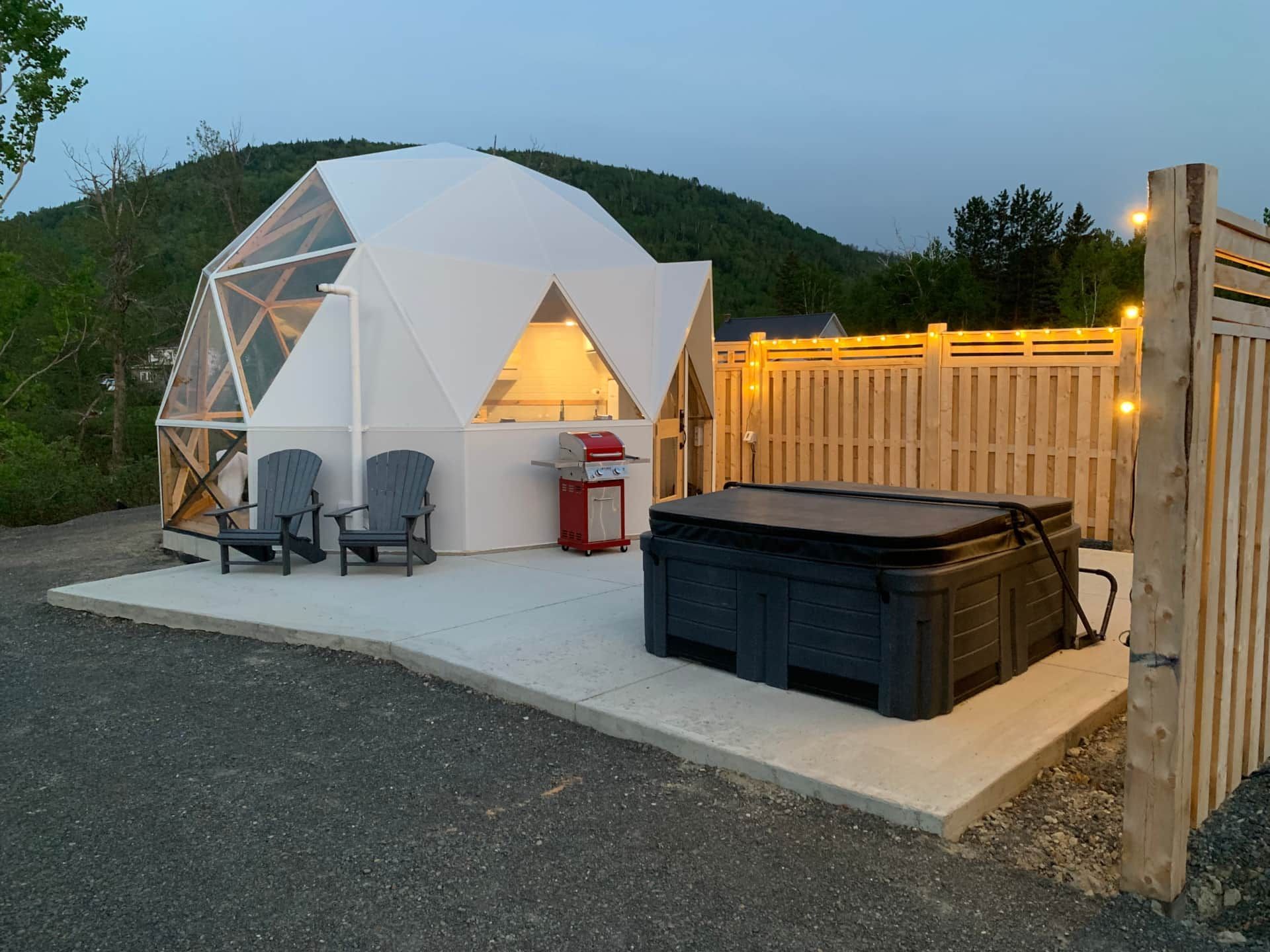 Witte geodetische dome van Appalaches Domes naast een verlicht houten hek, met een jacuzzi op een betonnen terras in de schemering.