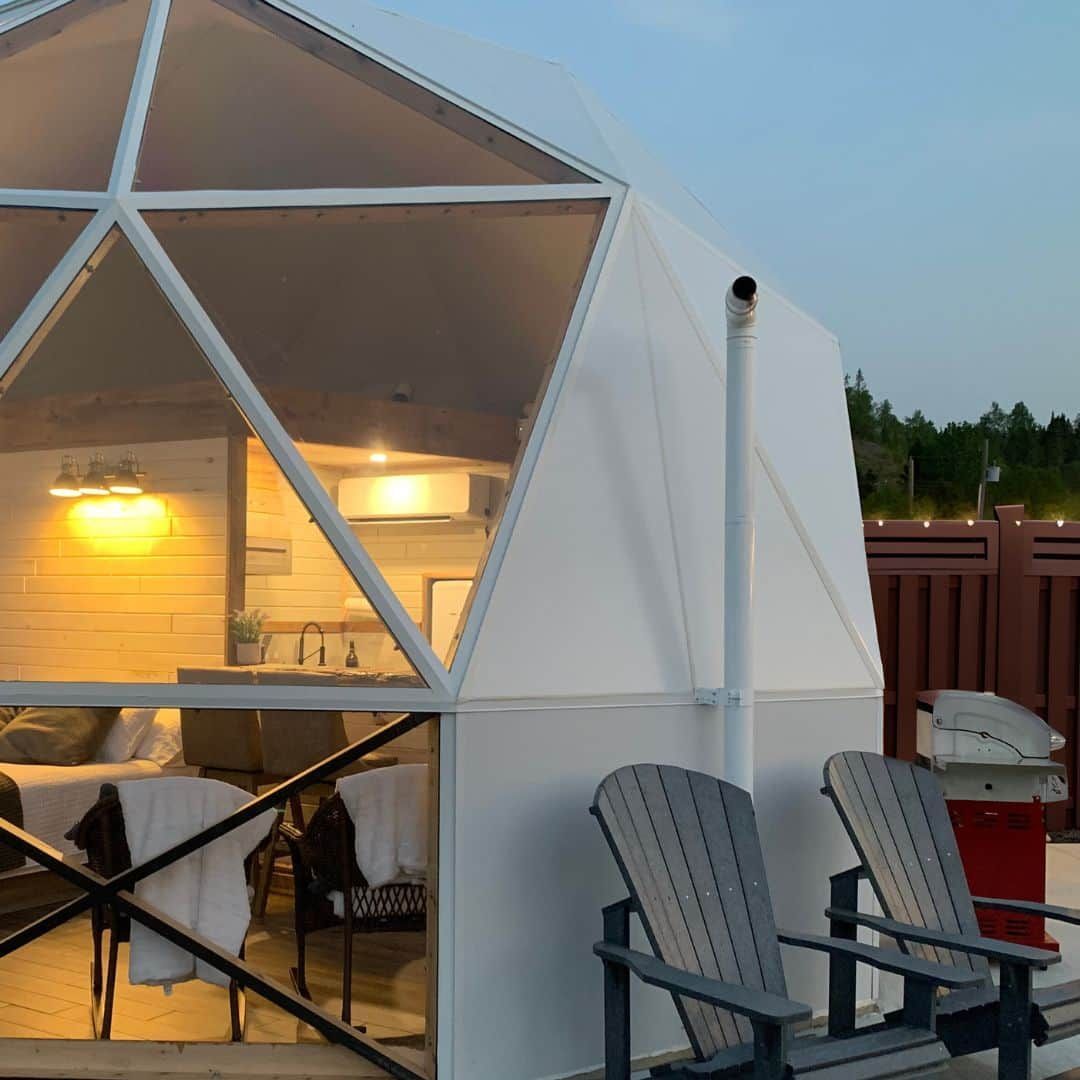 Witte geodetische dome van Appalaches Domes met terras, verlicht interieur, ligstoelen en een zonsondergang buiten.
