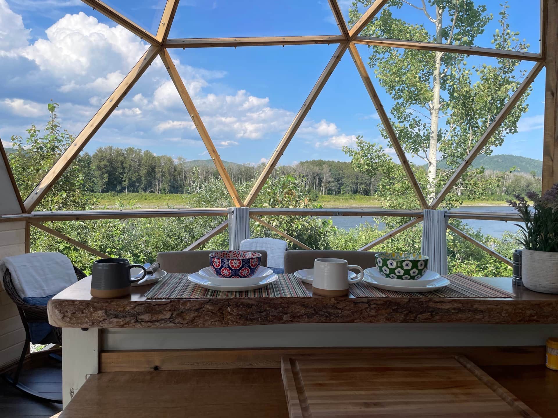Gedekte tafel aan een rivier, gezien door een dome van Appalaches Domes met daarachter een bosrijk landschap.
