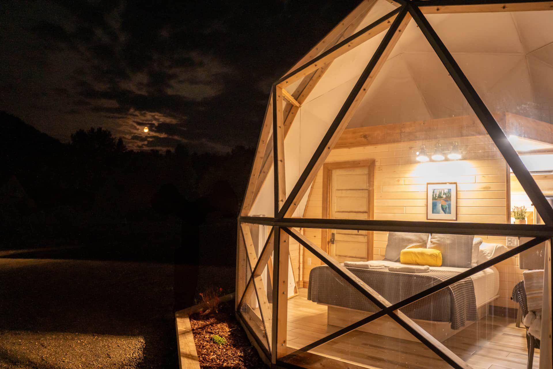 Een geodetische glampingtent van Appalaches Domes, 's nachts warm verlicht, staat tegen een donker landschap.