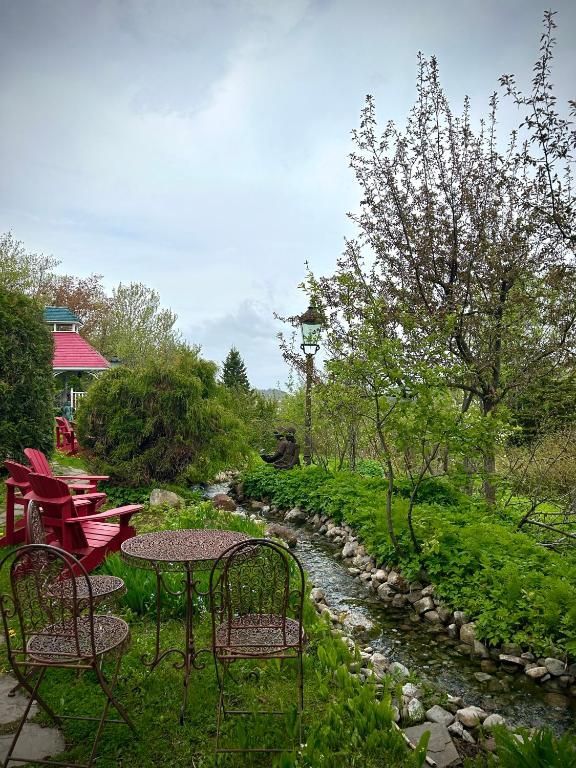 Tuinpatio met rode stoelen, een ronde tafel en een met stenen omzoomd beekje naast weelderig groen en bloeiende bomen.