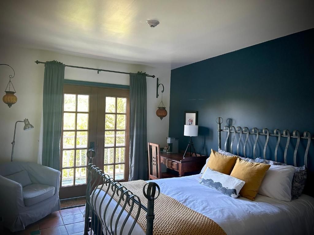 Gezellige slaapkamer met blauwe muren, een smeedijzeren bed, een grijze fauteuil en openslaande deuren met groene gordijnen.