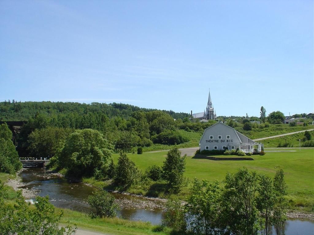 Een landelijke kerk en een wit huis aan een rivier in een groen landschap onder een blauwe hemel.