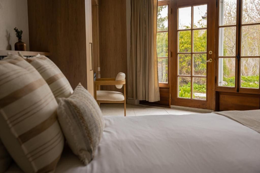 Gezellige slaapkamer met wit beddengoed, beige kussens, houten wanden en openslaande deuren naar een groene tuin.