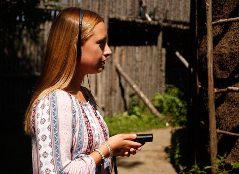 Vrouw met een telefoon in haar hand naast een rustieke houten schutting in het zonlicht.