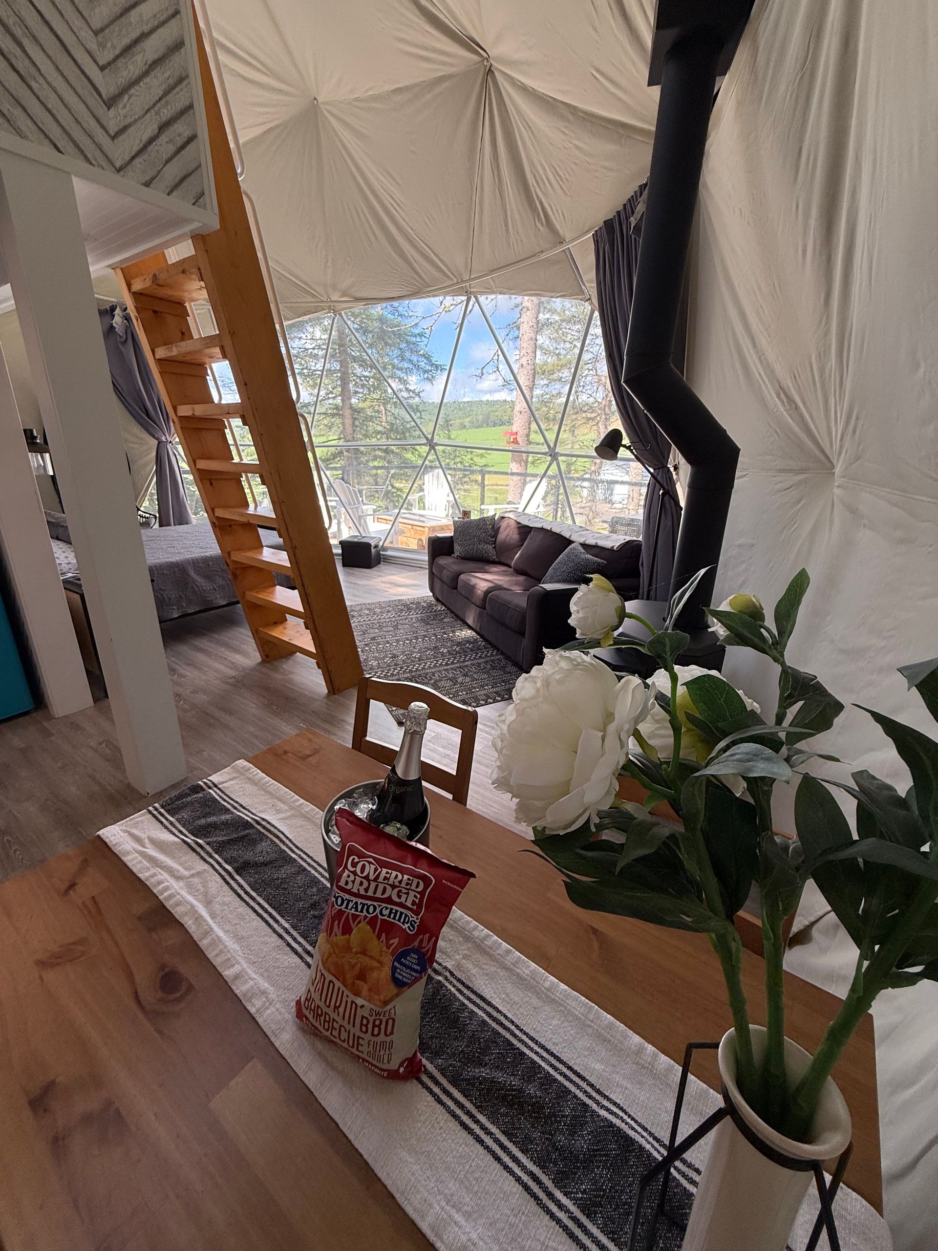 Gezellige woonkamer in een dome tent van Balsam Ridge Forest Domes met wit tentdoek, bank, ladderplank, planten en een salontafel met snacks en bloemen.