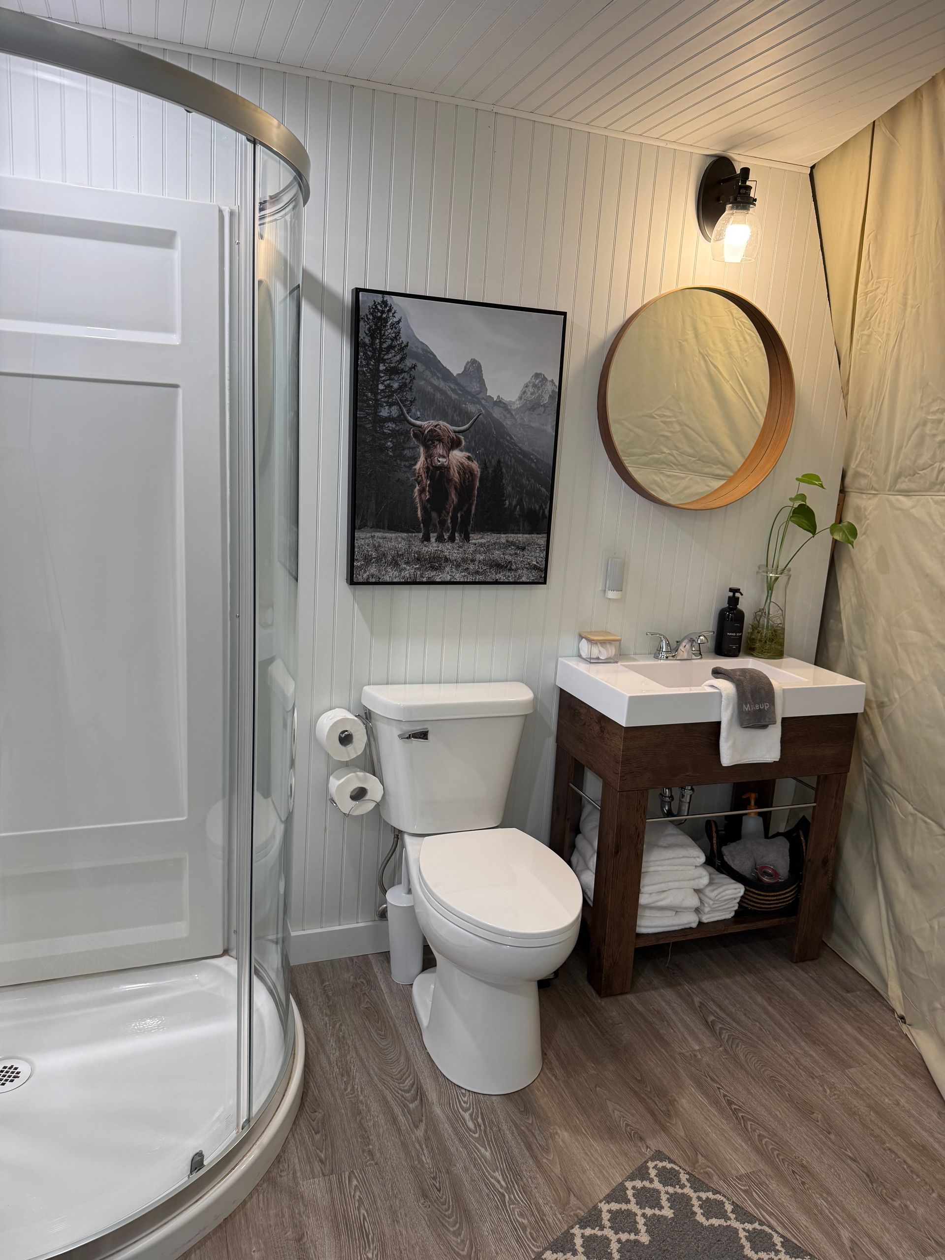 Badkamer in een dome tent van Balsam Ridge Forest Domes met douche, toilet, houten wastafelmeubel, ronde spiegel en muurdecoratie met bergmotief.