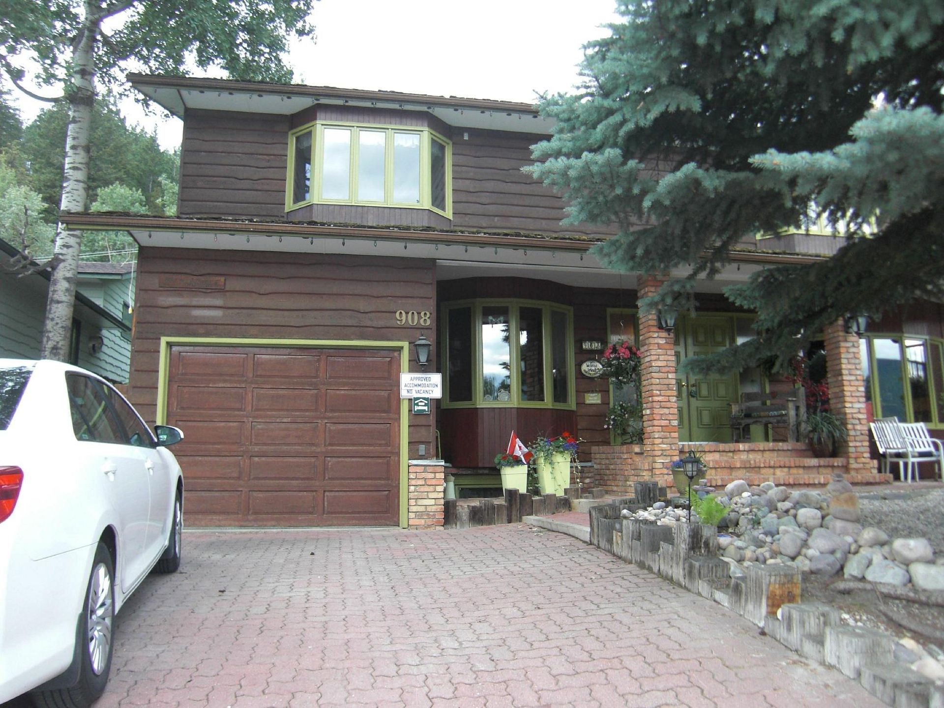 Bruin huis met twee verdiepingen, garage, bakstenen oprit en veranda, omgeven door bomen.