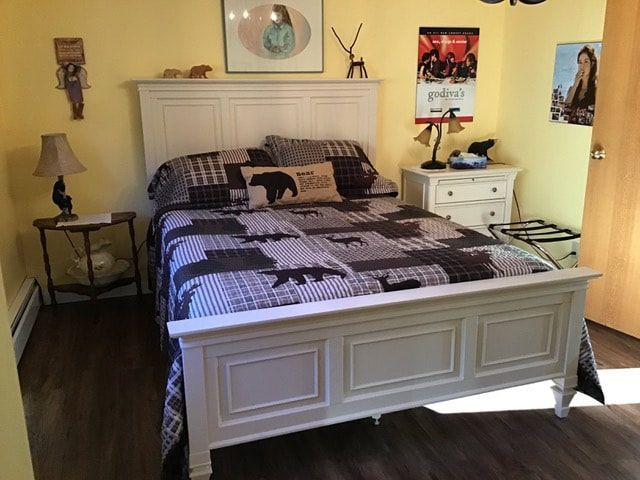 Wit bed met donker geruit beddengoed in een gele slaapkamer met nachtkastjes en wanddecoratie.