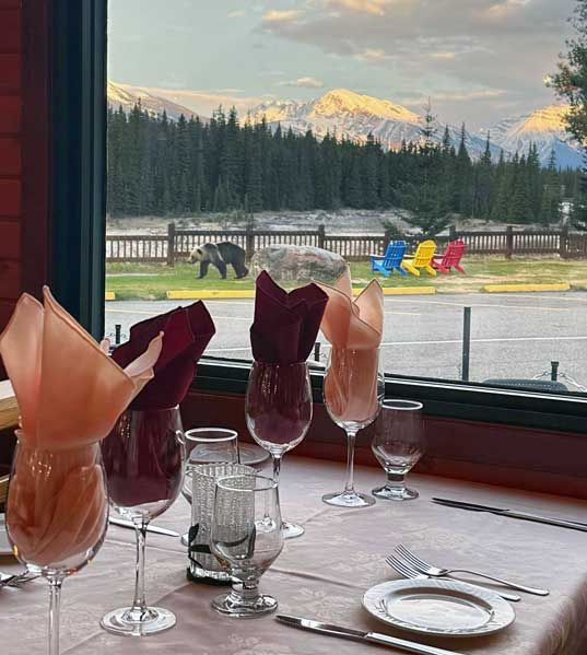 Eettafel bij een raam van Becker's Chalets Jasper met gevouwen servetten en glazen, met uitzicht op een rivier, bos en besneeuwde bergtoppen.