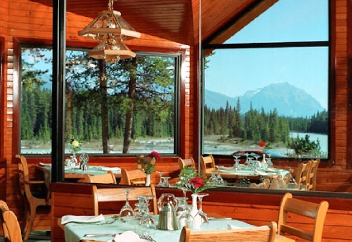 Gezellig restaurant van Becker's Chalets Jasper met grote ramen die uitkijken op een bos en de bergen in de verte, en een keurig gedekte tafel.