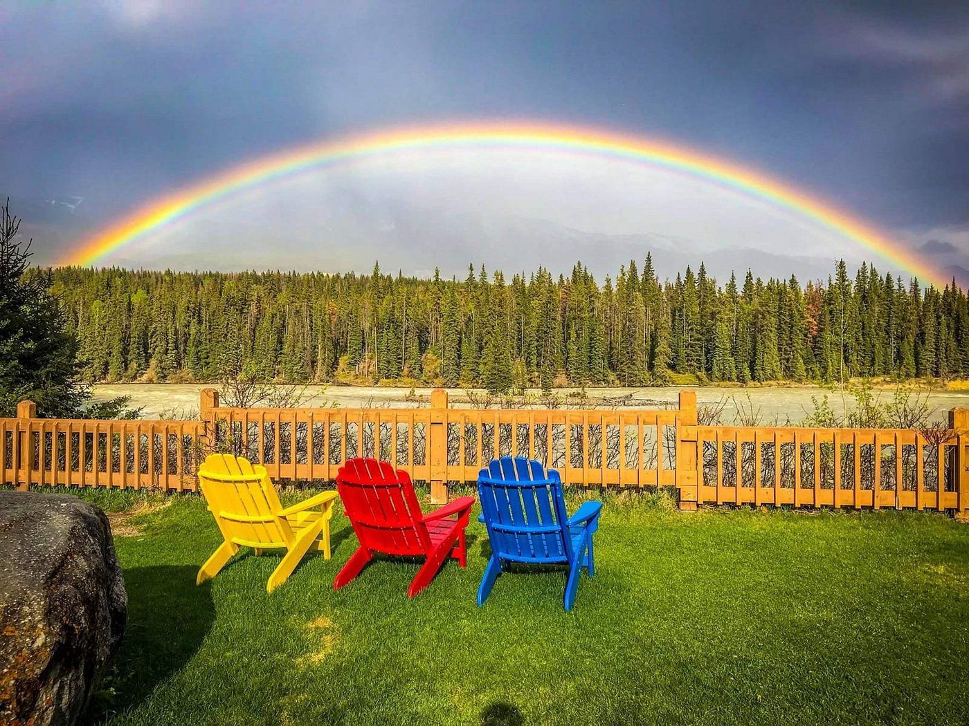 Een regenboog boven een achtertuin met gele, rode en blauwe stoelen op groen gras bij een houten hek te Becker's Chalets Jasper.