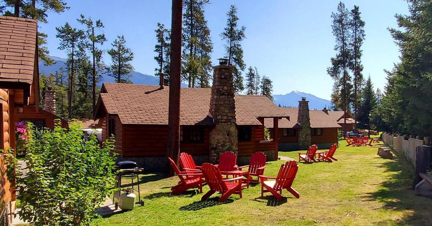 Rustieke hutten van Becker's Chalets te Jasper met rode Adirondack-stoelen op een zonnig gazon tussen dennenbomen en bergen.