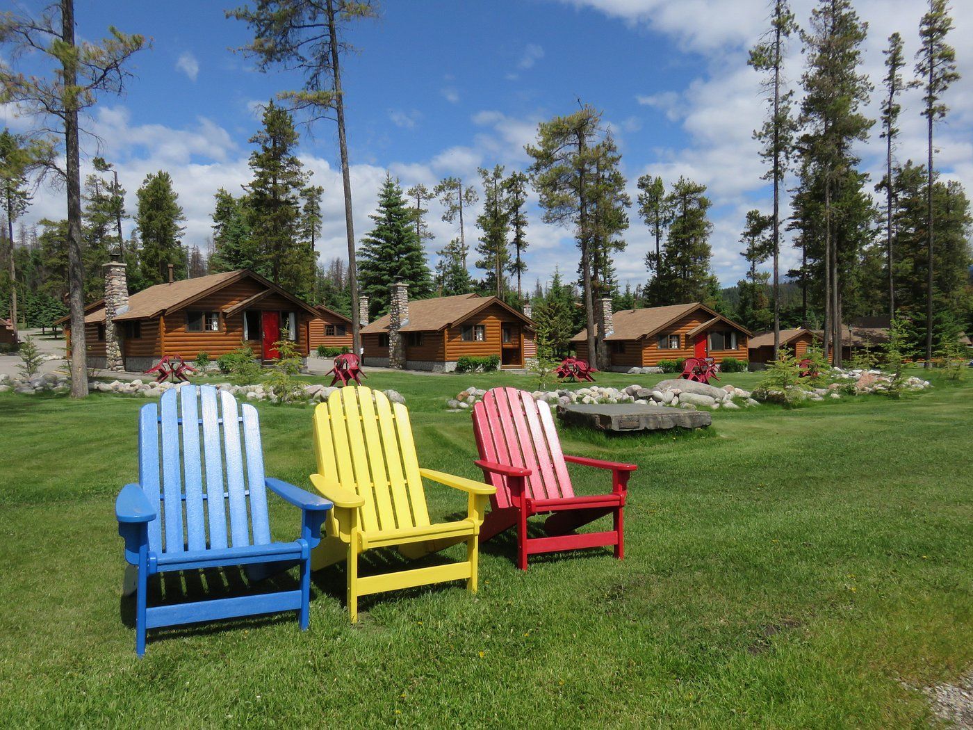 Kleurrijke Adirondack-stoelen op een grasveld bij rustieke hutten van Becker's Chalets Jasper en hoge dennenbomen onder een blauwe hemel.