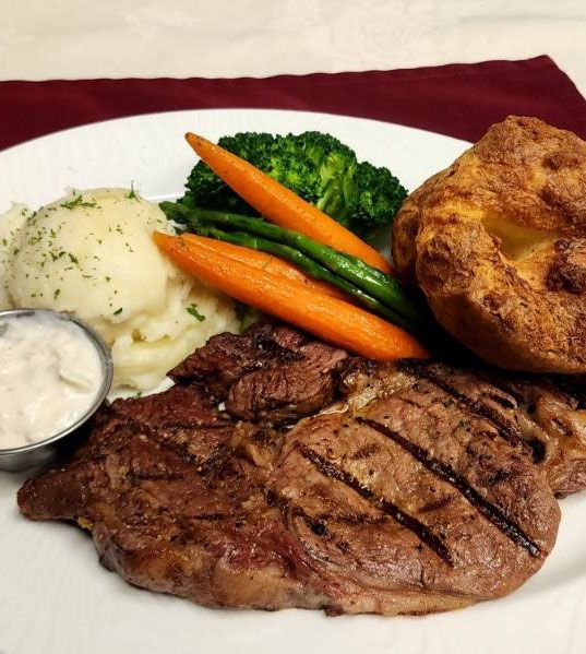 Gegrilde biefstuk met aardappelpuree, broccoli, wortelen en een broodje op een wit bord.