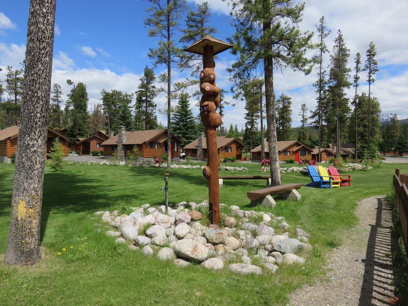 Hutten van Becker's Chalets Jasper met een gebeeldhouwde houten totempaal, een groen gazon, dennenbomen, hutten en kleurrijke speeltoestellen.