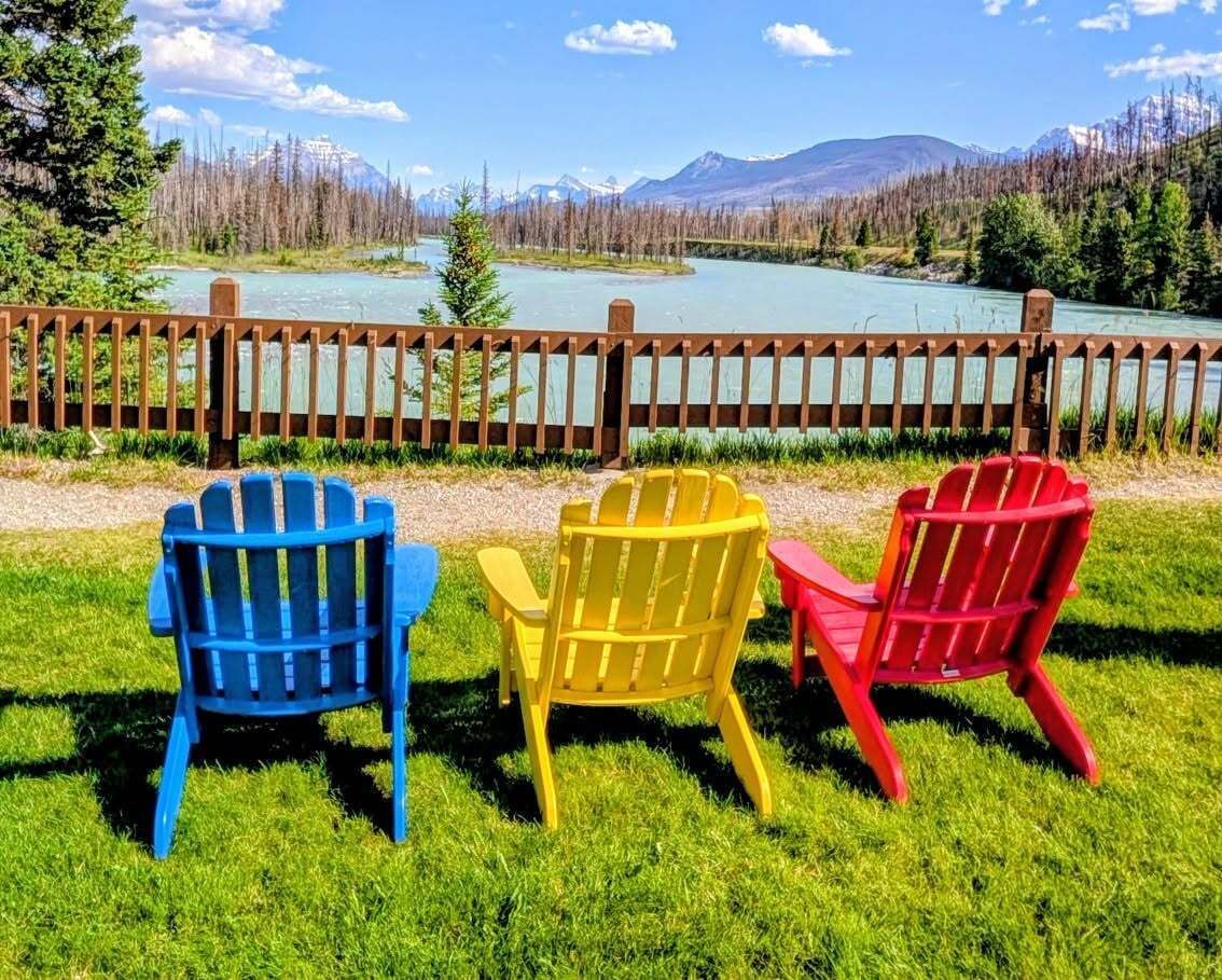 Drie kleurrijke Adirondack-stoelen van Becker's Chalets Jasper met uitzicht op een meer en bergen op een zonnige dag.