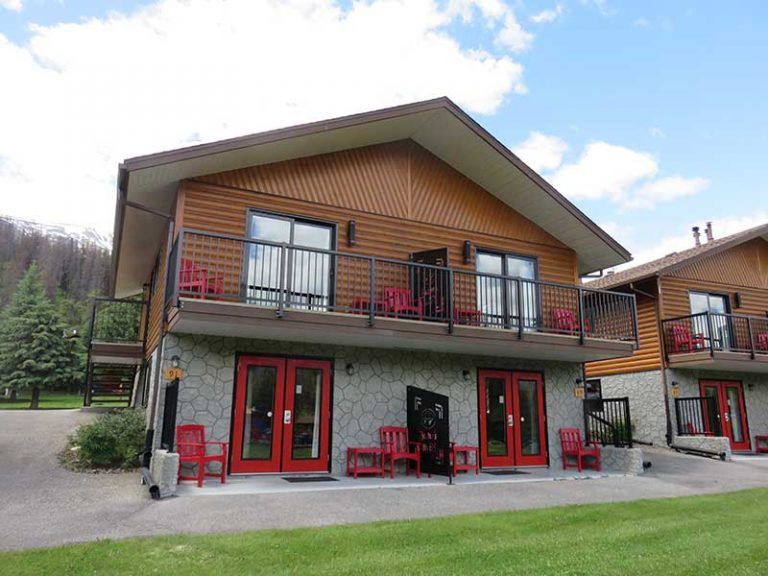 Een lodge van Becker's Chalets te Jasper met twee verdiepingen, rode deuren, een balkon en een terras in een groene bergachtige omgeving.