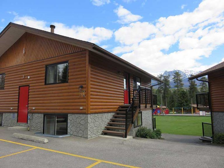 Een houten gebouw in blokhutstijl van Becker's Chalets te Jasper met een rode deur, een buitentrap en een stenen fundering, onder een gedeeltelijk bewolkte hemel.