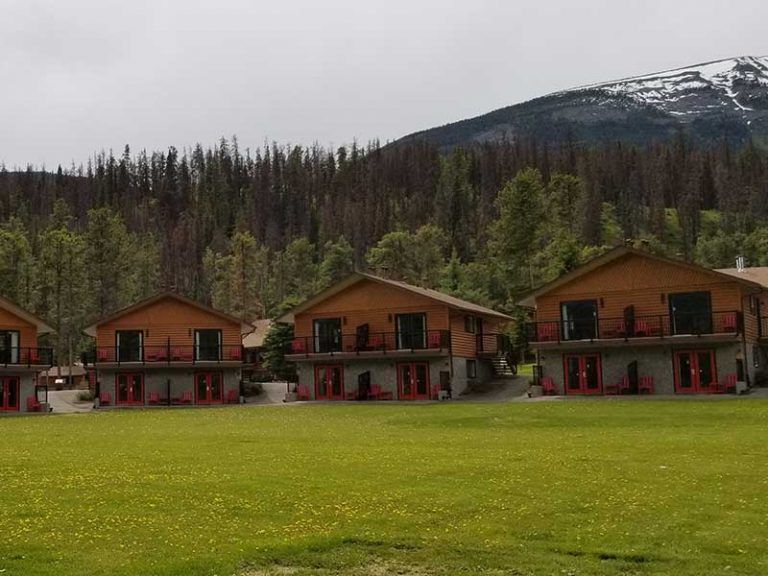 Een rij blokhutten van Becker's Chalets te Jasper in een groen veld met bos en besneeuwde bergtoppen op de achtergrond.