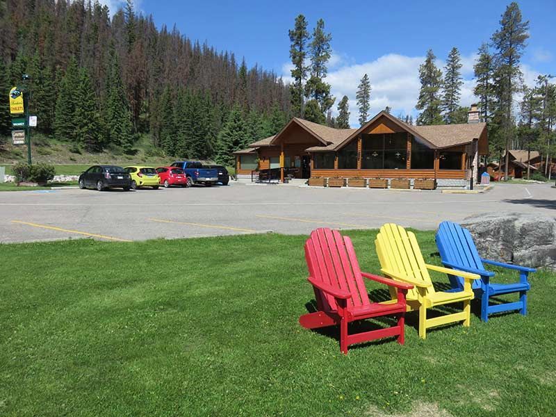 Kleurrijke Adirondack-stoelen op een gazon bij Becker's Chalets Jasper, met geparkeerde auto's en dennenbomen op de achtergrond.
