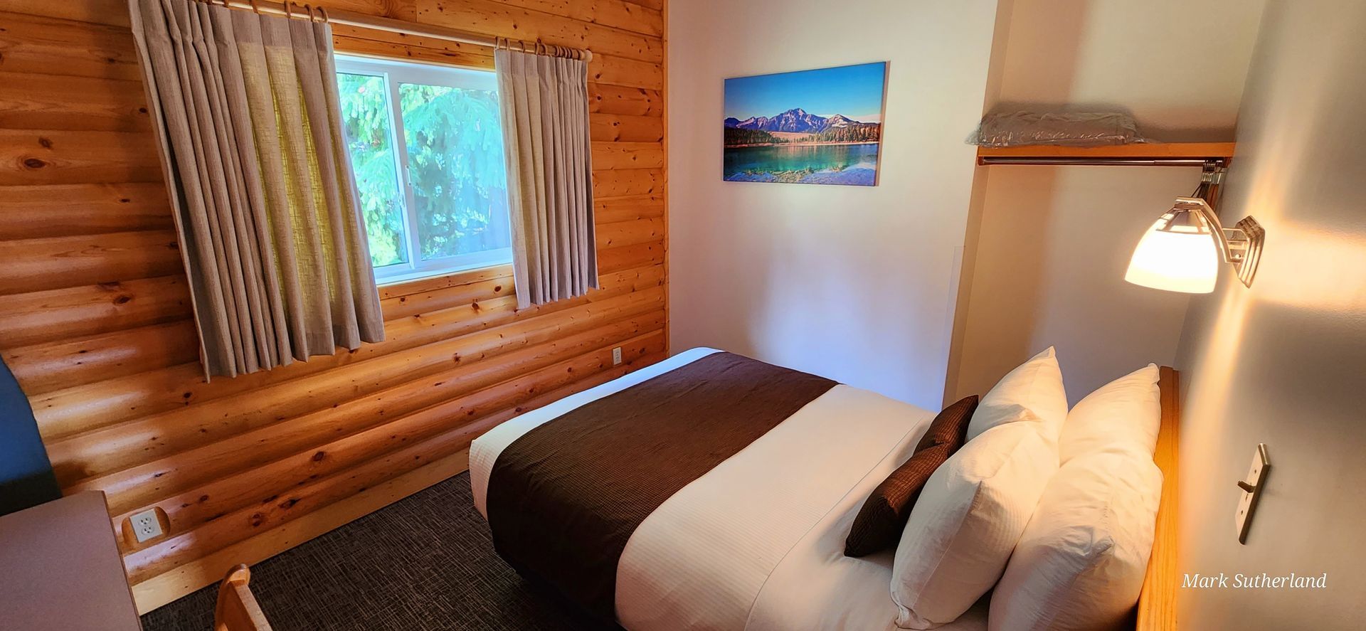 Gezellige slaapkamer in een blokhut met twee bedden, een wandlamp, een raam en een kunstwerk met een berglandschap.