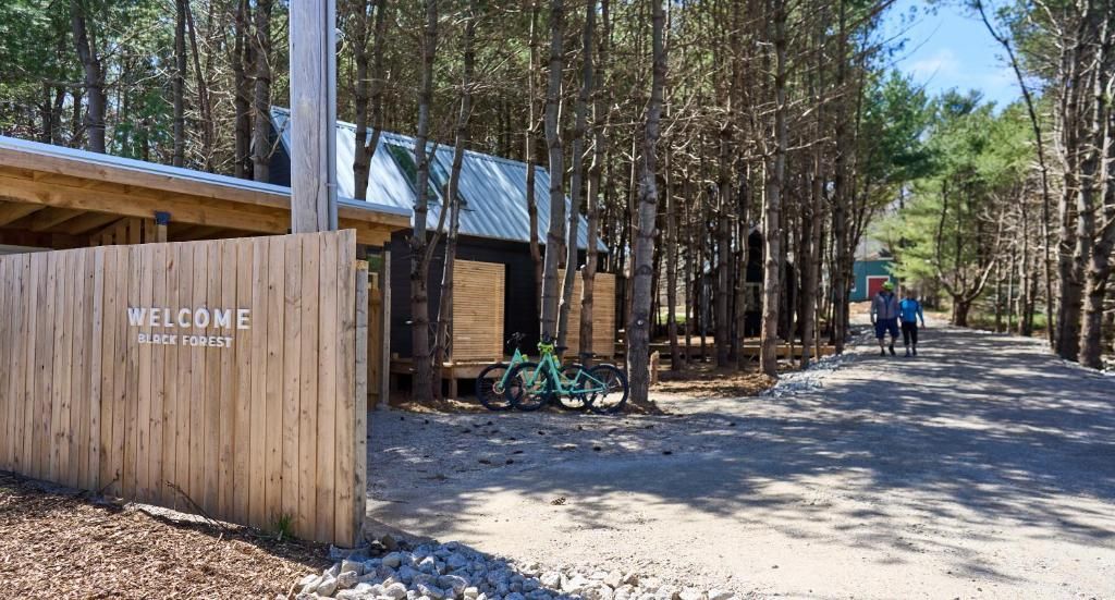 Een houten toegangspoort van Black Forrest Cabins, Mahone Bay naast een zandpad, met mensen die in de verte wandelen.
