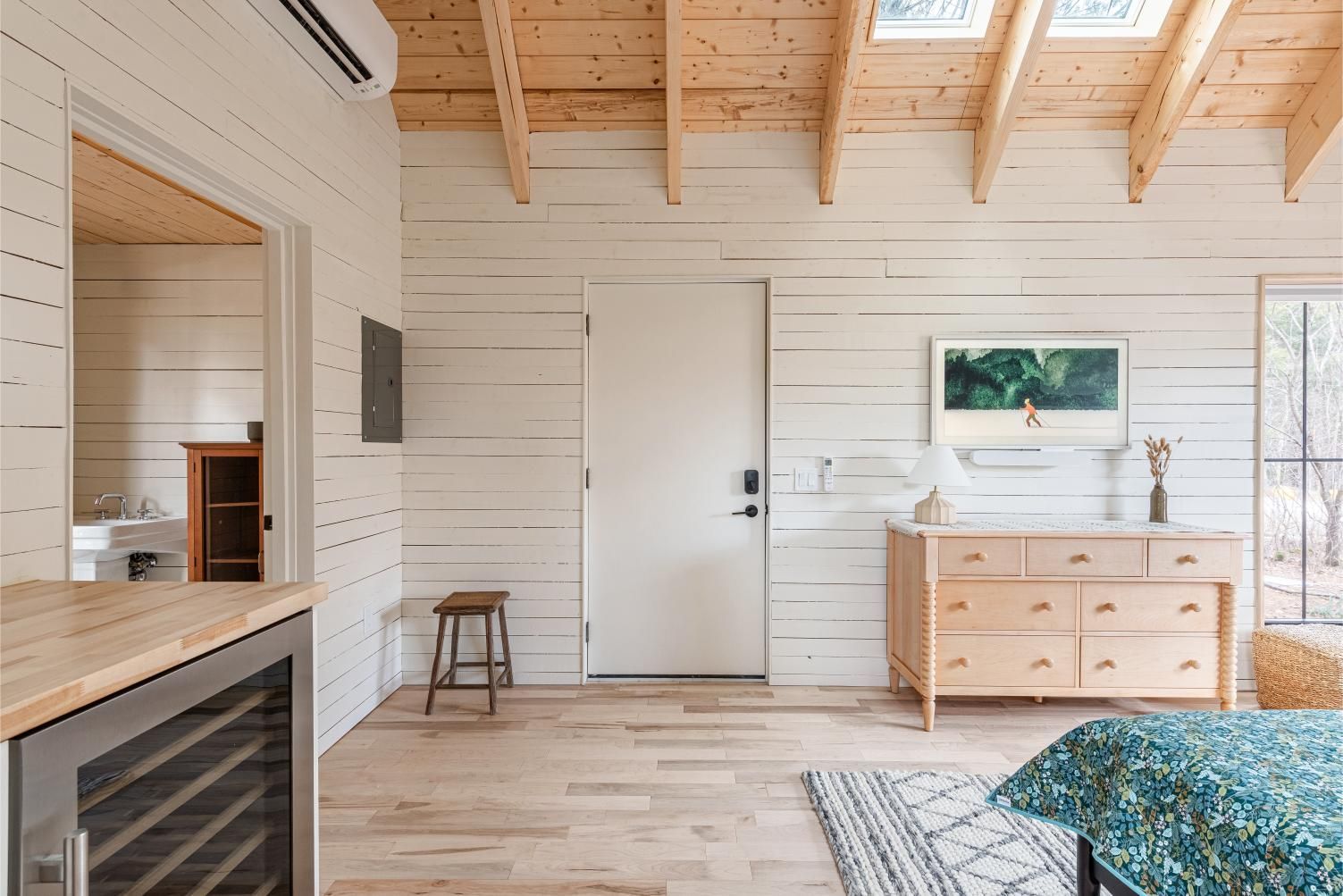 Lichte, rustieke slaapkamer van Black Forrest Cabins, Mahone Bay met witte houten lambrisering, houten plafondbalken, een commode en een schoorsteenmantel.