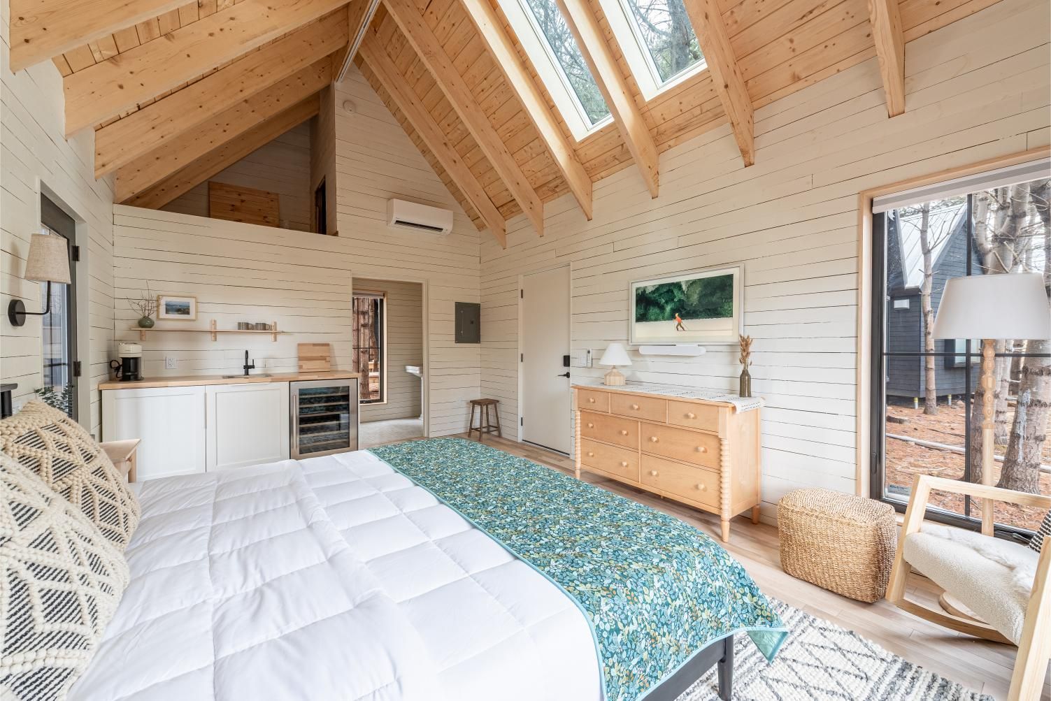 Gezellige loftslaapkamer van Black Forrest Cabins, Mahone Bay met houten balken, witte muren, bed, commode, kitchenette en openslaande deuren naar het balkon.