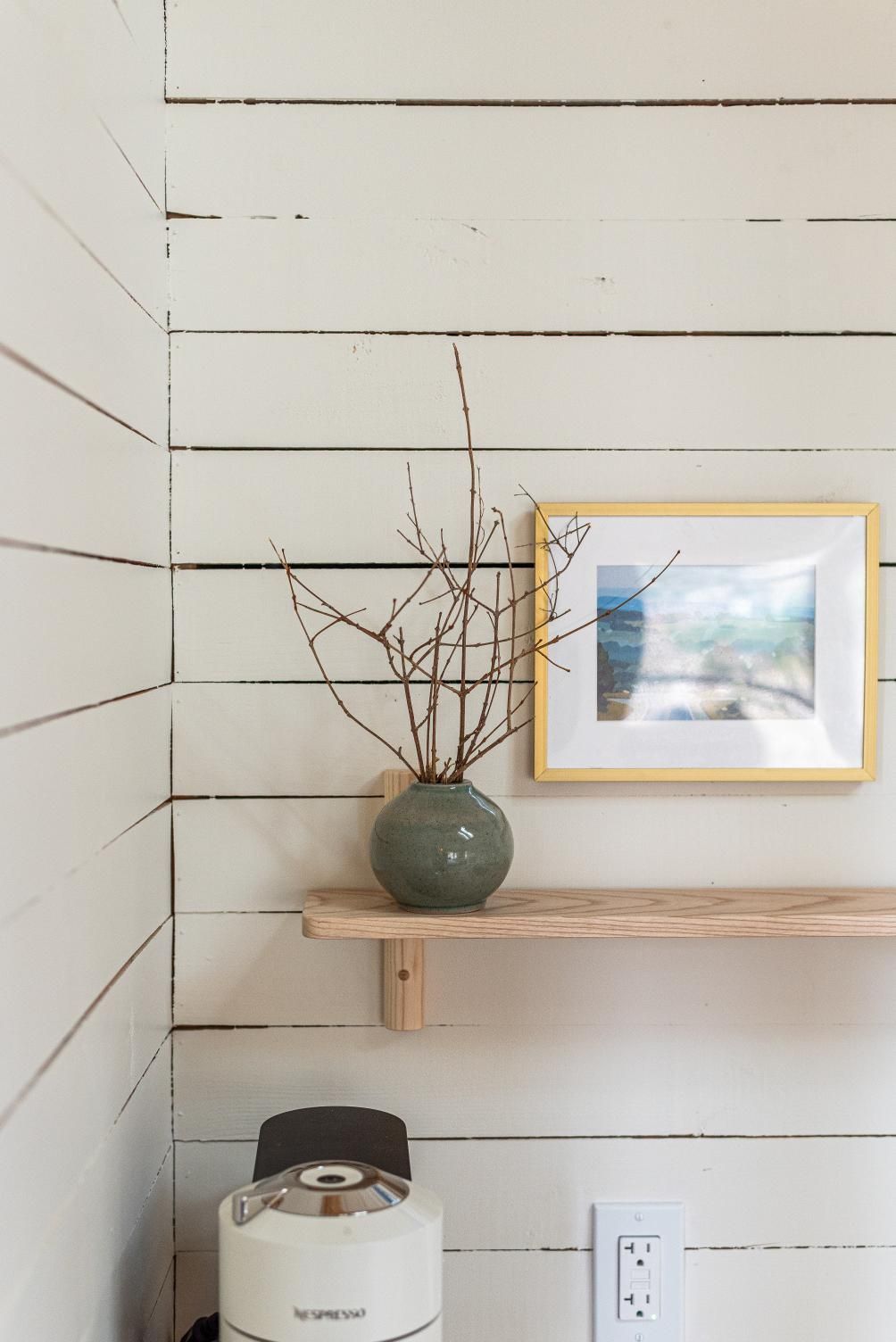 Minimalistische wandplank met een groene vaas met takken en een ingelijste landschapsschilderij op een witte lambrisering in een cabin van van Black Forrest Cabins, Mahone Bay.