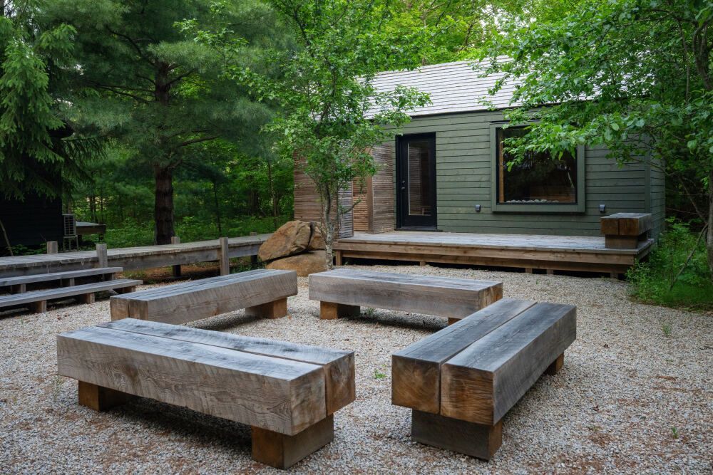 Een grindterras in de buitenlucht met houten zitbanken en een klein, modern tuinhuisje, omgeven door bomen bij Black Forrest Cabins, Mahone Bay.