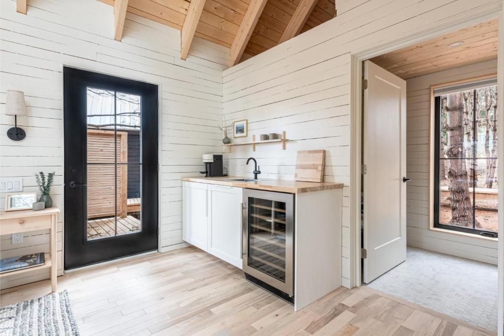 Lichte, kleine keuken met witte kastjes, zwarte deur, houten balken en een kleine wijnkoelkast, die uitkomt op een gang in een cabin van Black Forrest Cabins, Mahone Bay.