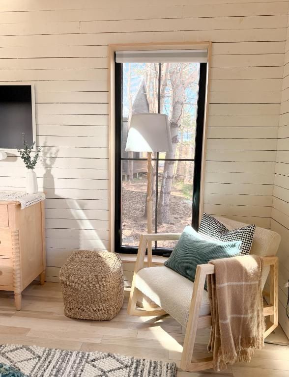 Gezellig, zonnig leeshoekje van Black Forrest Cabins, Mahone Bay met stoel, geweven poef, staande lamp en uitzicht op de bomen vanuit het raam.