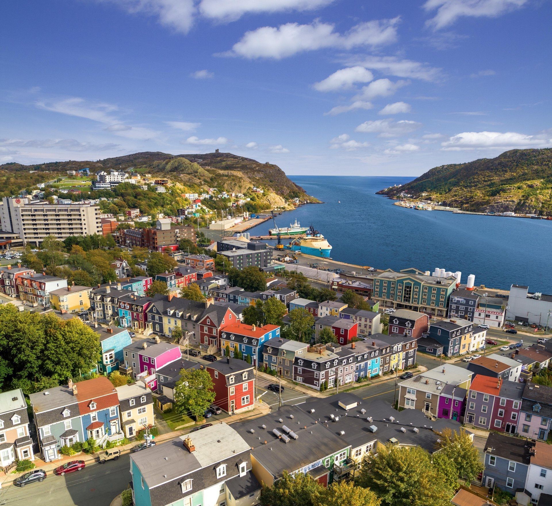 Het kleurrijke St. John's, de hoofdstad van Newfoundland, aan het water met heuvels, een haven en een blauwe baai onder een gedeeltelijk bewolkte hemel.