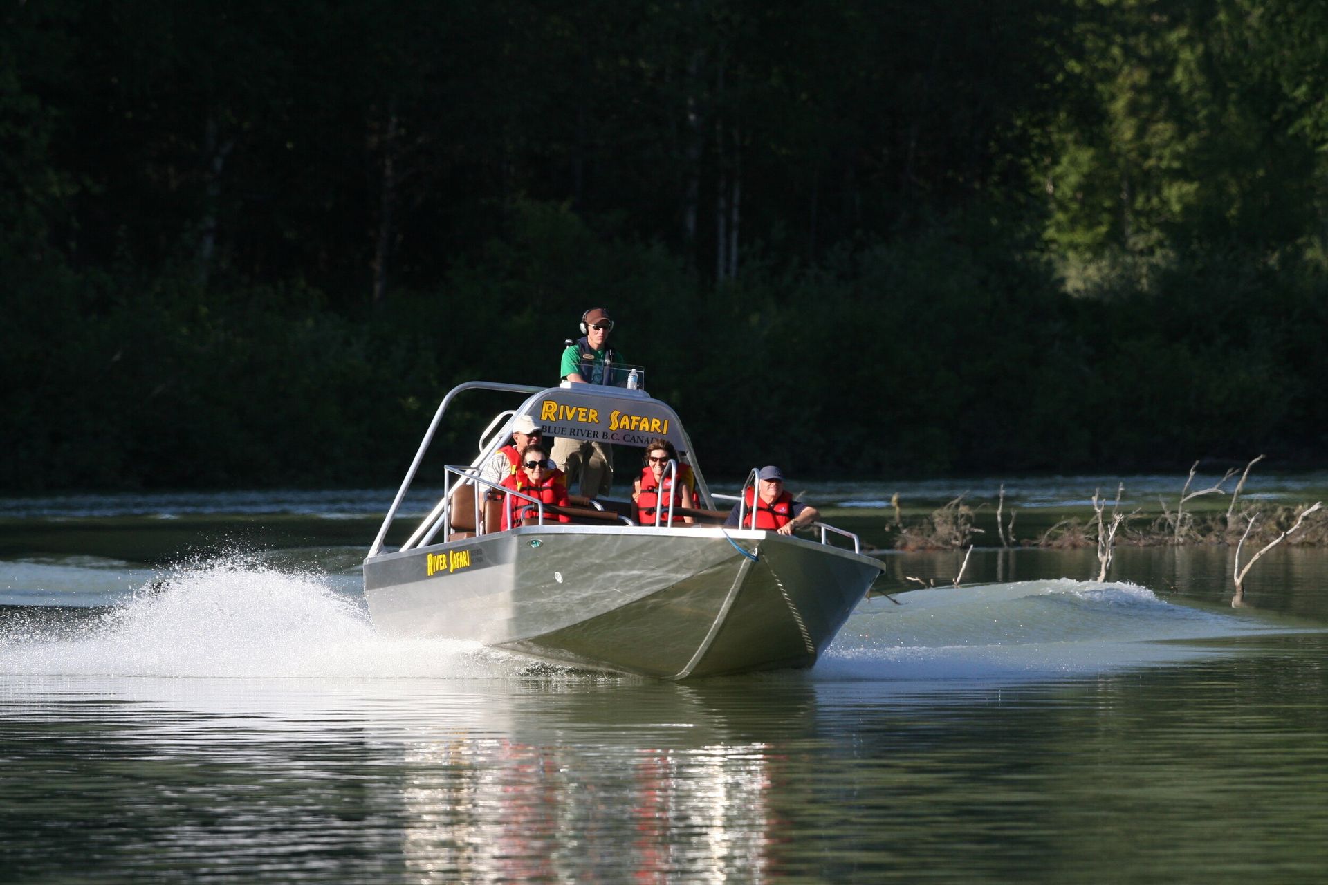 Een speedboot raast over een meer en spat water omhoog, met passagiers aan boord nabij een beboste oever.
