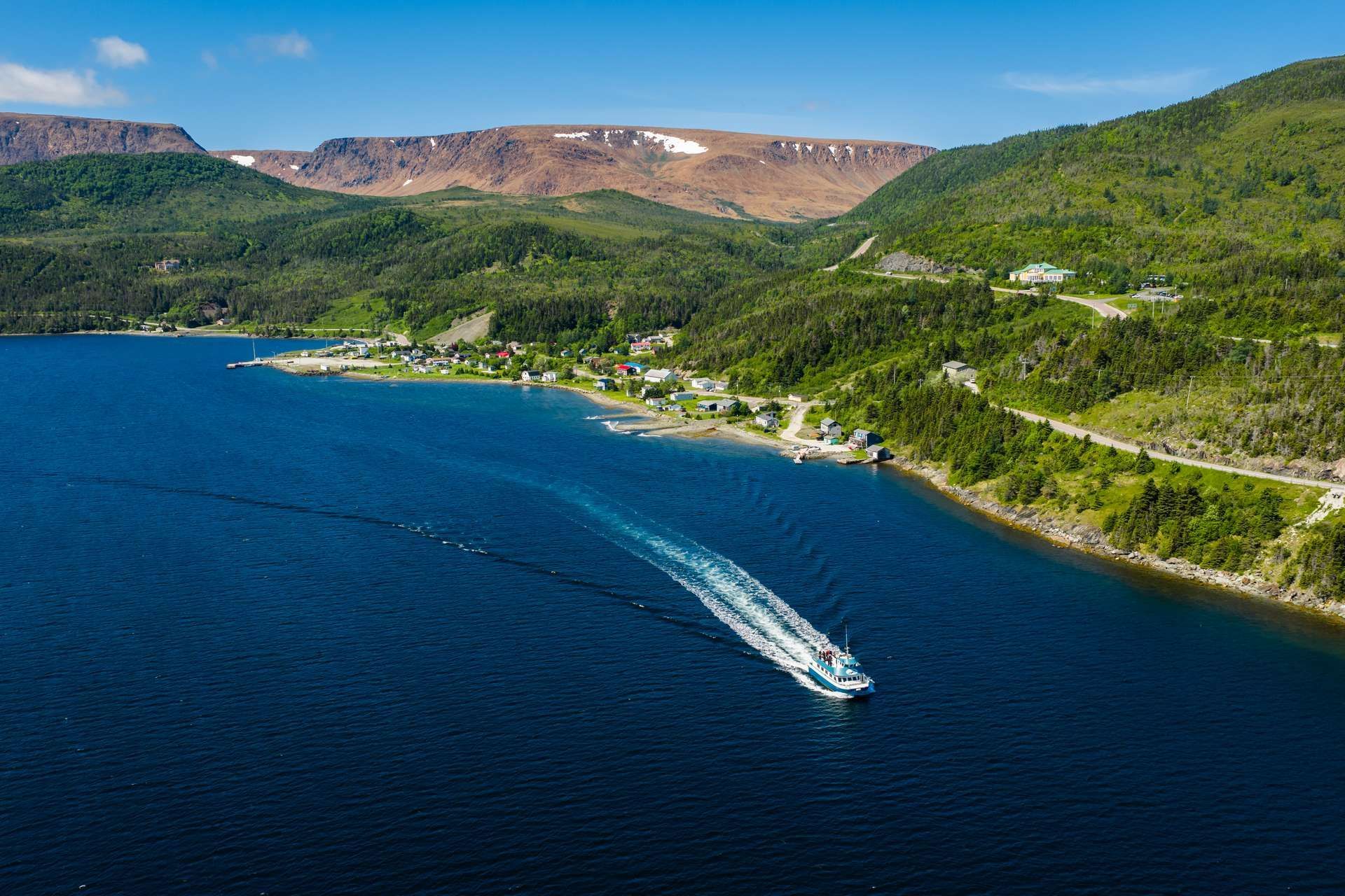 Een speedboot vaart door een diepblauwe baai, langs groene heuvels en rotsachtige bergen onder een heldere hemel.