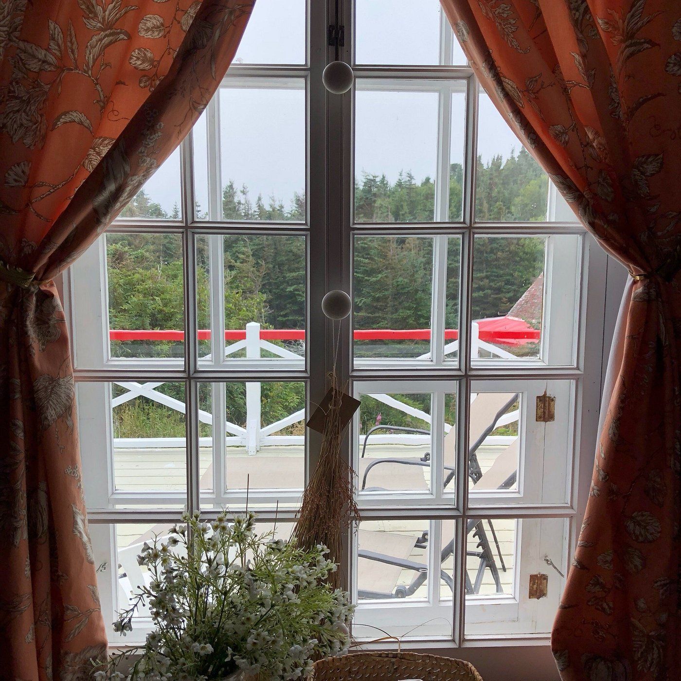 Rode gordijnen omlijsten een raam met uitzicht op een balkon, groene bomen en een parasol buiten.