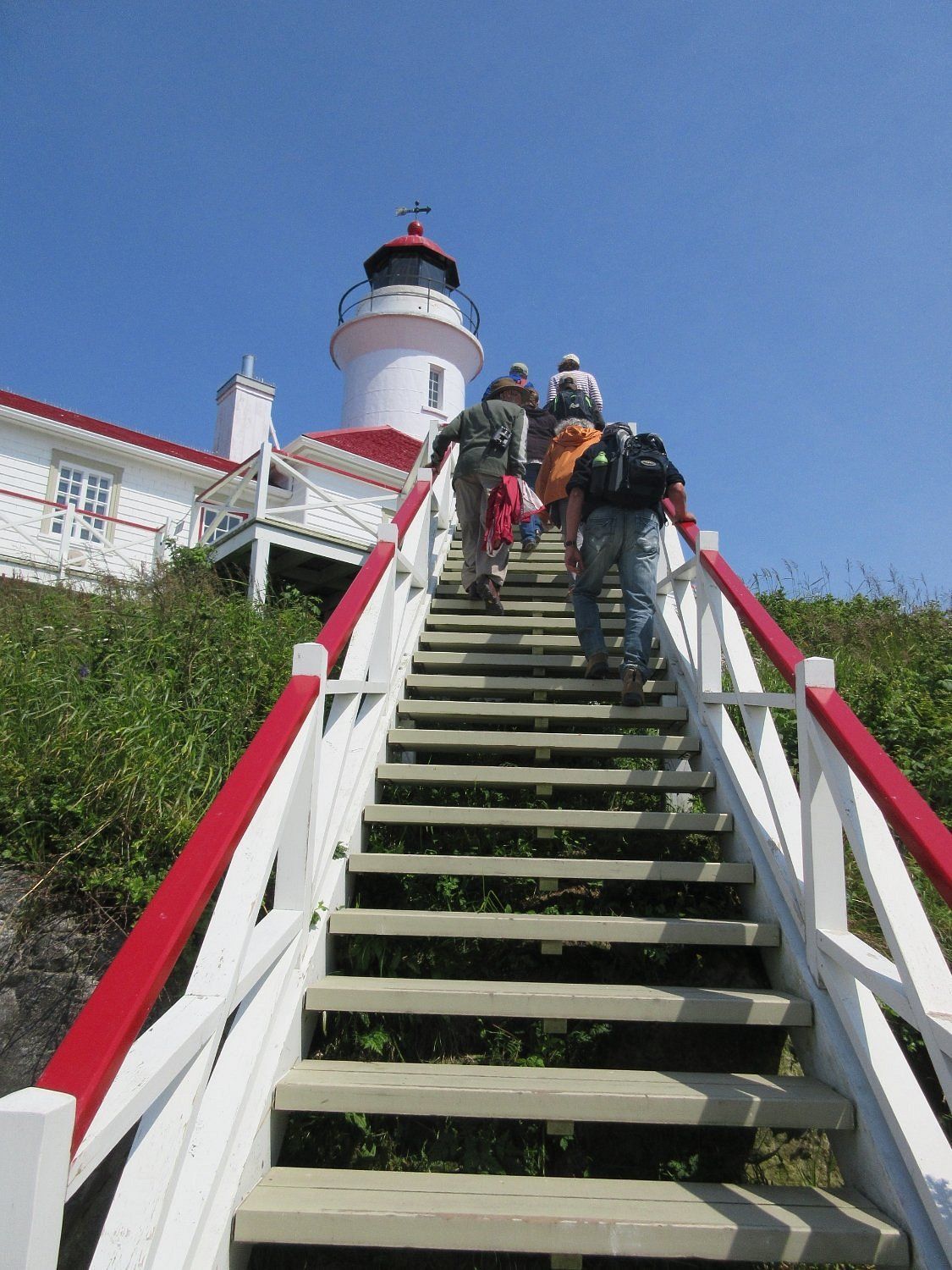 Mensen beklimmen steile wit-rode trappen naar een vuurtoren onder een helderblauwe hemel.