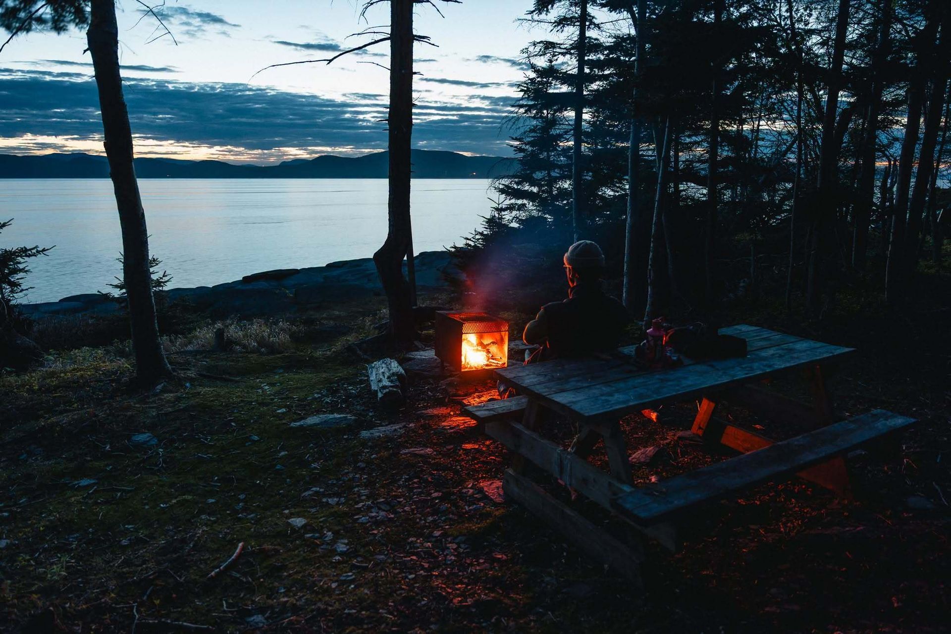 Een kampeerplek aan een meer in de schemering, met een brandend vuur naast een picknicktafel en silhouetten van bomen.