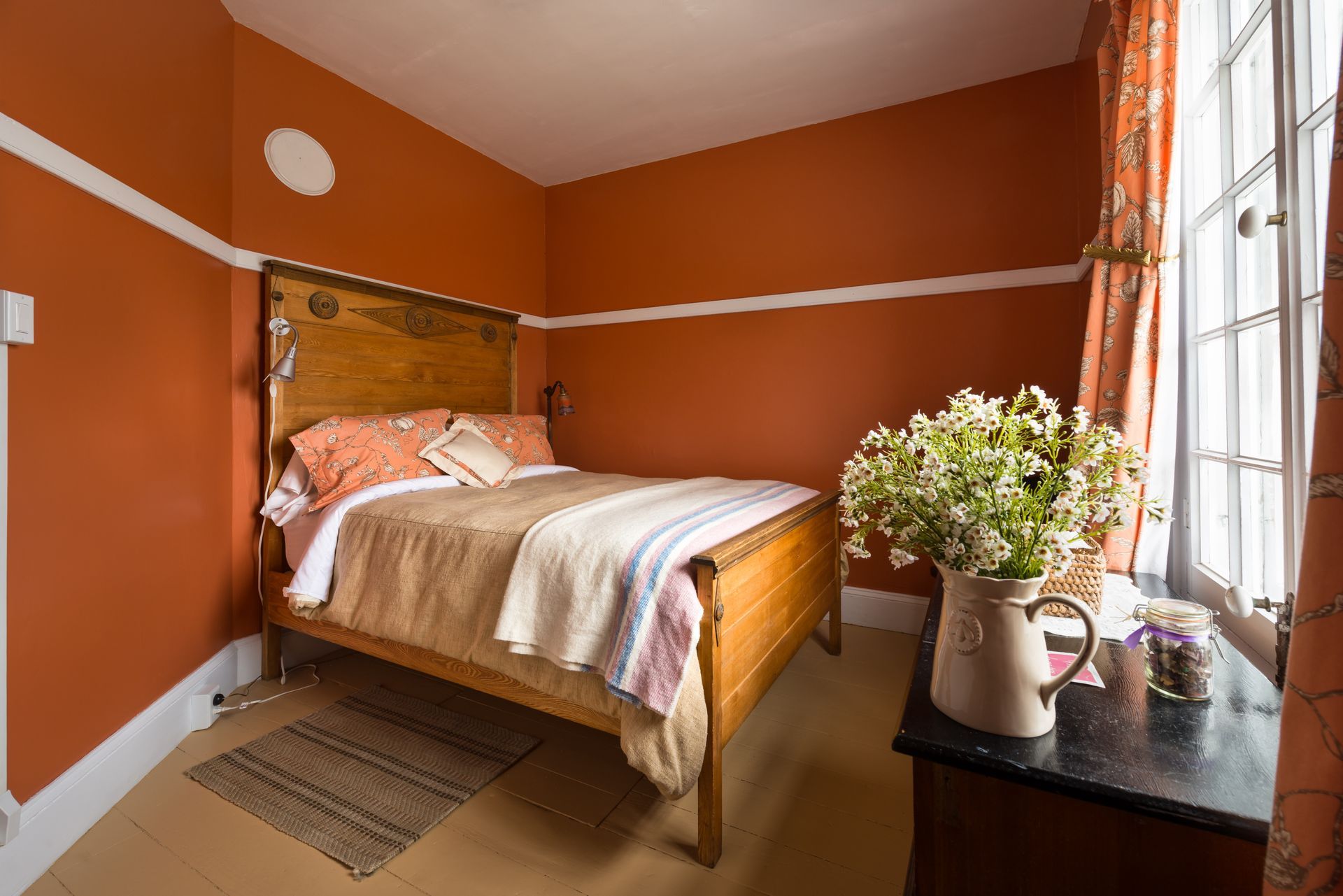 Gezellige slaapkamer met oranje muren, een houten bed en bloemen bij een zonnig raam.