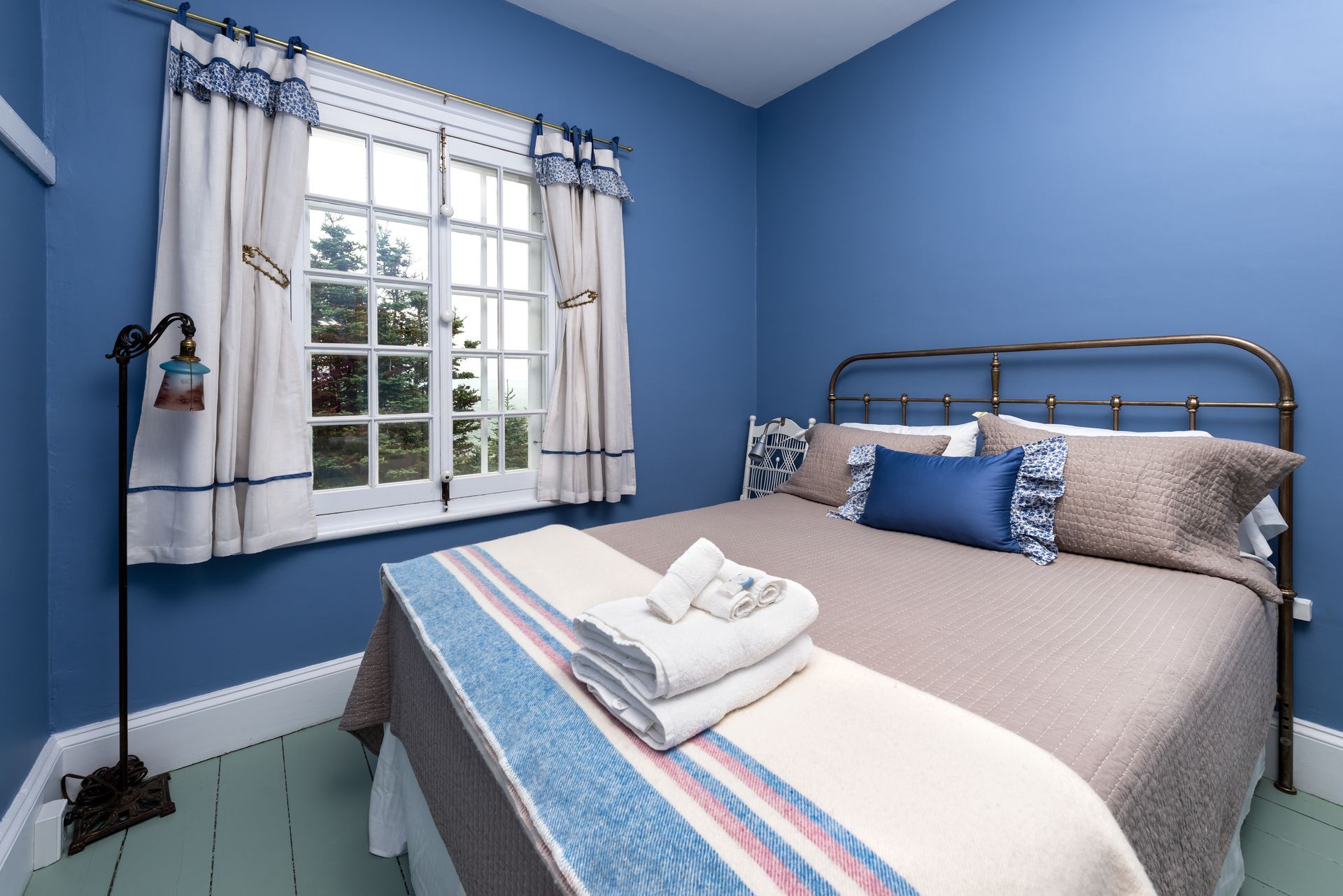 Blauwe slaapkamer met een wit bed, een messing hoofdbord en een raam met gordijnen.
