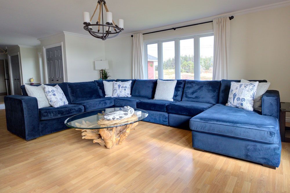 Lichte woonkamer met een donkerblauwe hoekbank, glazen salontafel, houten vloeren en grote ramen.