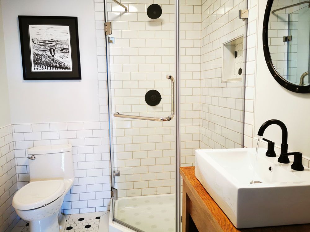 Moderne badkamer met witte tegels, toilet, glazen douchecabine en zwarte kranen naast een houten wastafelmeubel.