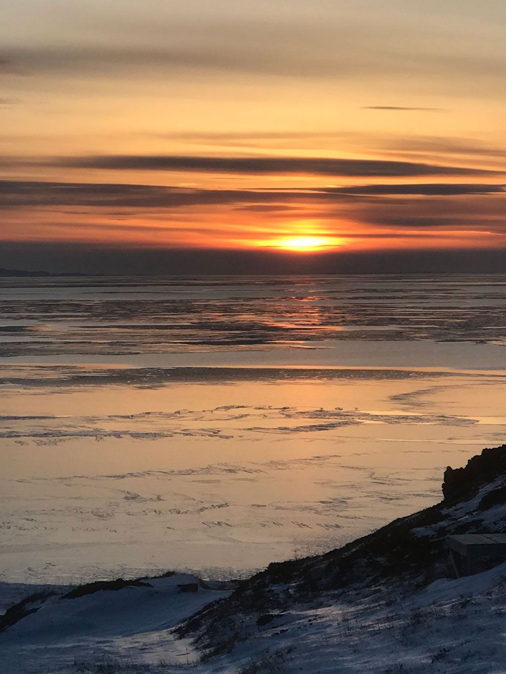 Zonsondergang boven een besneeuwde kustlijn met een oranje lucht die weerspiegelt in het ijskoude water.